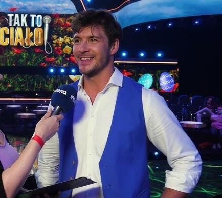Mikołaj Roznerski na planie filmowym, w dynamicznej pozie, oddający charakter jednej ze swoich ról.