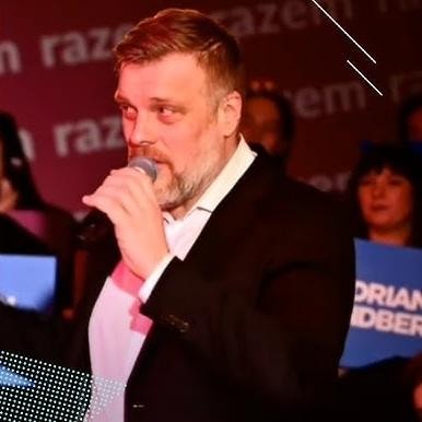 Adrian Zandberg, polski polityk, uśmiechnięty i patrzący prosto w obiektyw, w swobodnym, ale eleganckim ubraniu, na tle delikatnie rozmytego, ciepłego wnętrza, emanujący serdecznością i inteligencją.