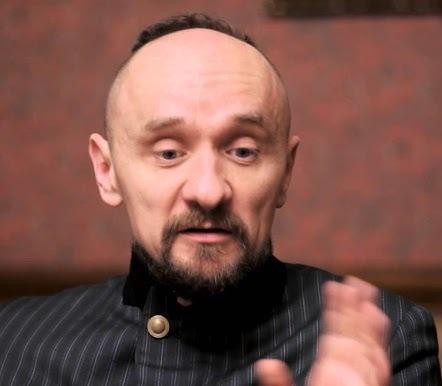 Jan Klata w dynamicznej scenie teatralnej, gestykulujący na tle rozmazanych elementów scenografii i postaci, oddający energię jego reżyserskiej pracy.