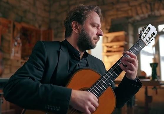 Marcin Dylla z gitarą na scenie sali koncertowej, widziany z perspektywy widowni, kłaniający się publiczności.