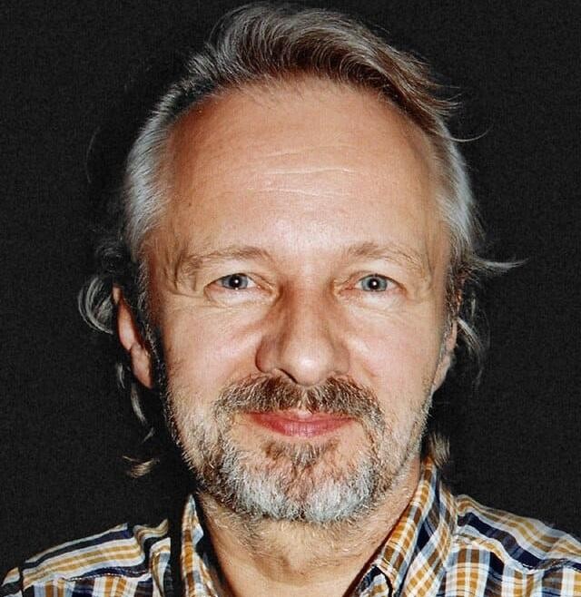 Krzysztof Stroiński uśmiechający się subtelnie, w ujęciu z bliska, na rozmytym tle, sugerującym plan filmowy lub ulicę.