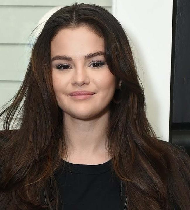 Selena Gomez na scenie lub planie zdjęciowym, prezentująca się pewnie i profesjonalnie, z rozmytym, artystycznym tłem.