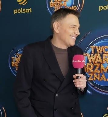 Krzysztof Ibisz w studiu telewizyjnym, z mikrofonem, w tle elementy scenografii programu.